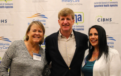 three white people (two women on each side of a man) standing in front of a press backdrop