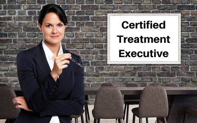 a white woman in a business suit in front of a table and a white board. Text on whiteboard reads 'Certified Treatment Executive'