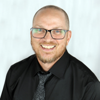 a white man with balding dark hair and a light brown beard wearing black glasses and a black button-up shirt and tie