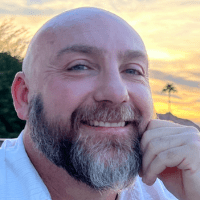 a bald middle-aged white man with a grayish-black beard, his left hand on his cheek, wearing a light blue shirt