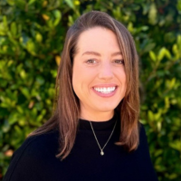a white woman with long brown hair, wearing a black turtleneck and a silver necklace, standing in front of some greenery