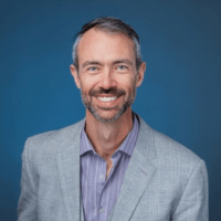 an older white man with a scruffy beard and mustache wearing a gray blazer with a light purple button up