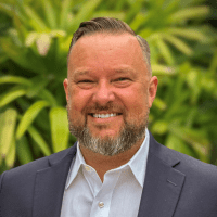 a middle-aged white man with a graying-black small beard, wearing a gray suit with a button-up white shirt underneath