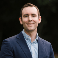 a young white man with short brown hair, wearing a dark blue suit jacket with a light blue button-up shirt underneath