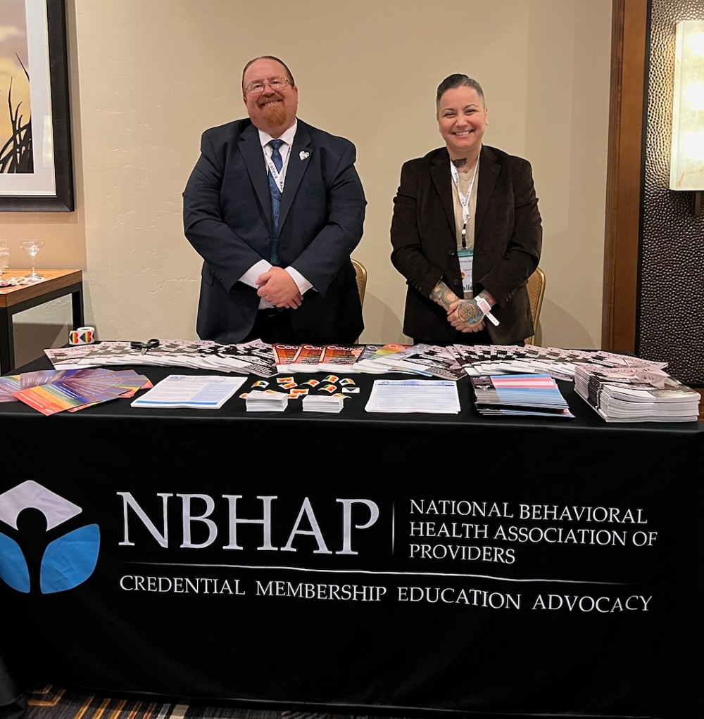 two people in suits standing behind a table with the NBHAP logo on it and lots of marketing materials on it. 