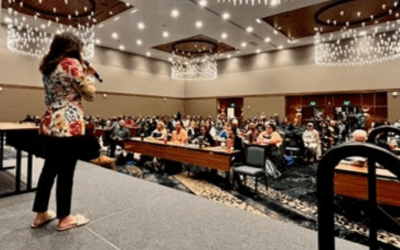 a woman stands on a stage with a microphone in her hand, with audience at desks