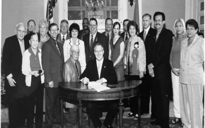 a black and white photo of several people surrounding a table, the center man signing a paper