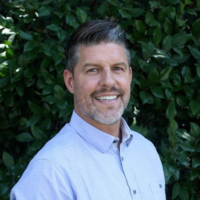 a Latinx man with black hair with a streak of white and a beard wearing a light blue button up shirt