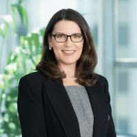 a white woman with shoulder-length brownish/black hair wearing glasses, a grey top, and a black suit jacket