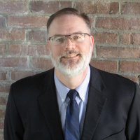 a white man with brown hair, a grey beard, and glasses wearing a dark suit on the right