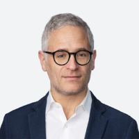 a white man with short grey hair, wearing black glasses and a dark blue blazer over a white top