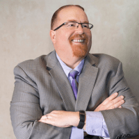 a middle-aged white man with red hair and a goatee wearing glasses and a light gray suit with a light purple shirt with a darker purple tie. He stands with his hands crossed, grinning
