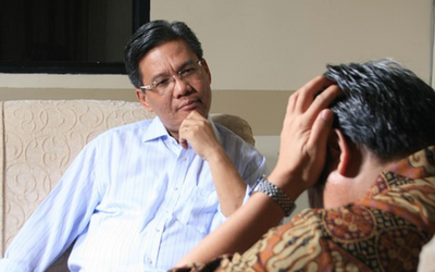 an Asian man with glasses and a light blue shirt, hand on chin looking toughtfully on a couch, looks at another man, hands on his head as if stressed