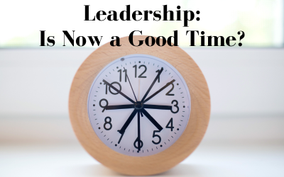 Brown wooden round analog clock with multiple hands indicating the time. Text reads, 'Leadership: Is Now a Good Time?'