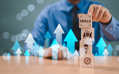 a white man in a light blue business button-up top with a dark blue sky sits at a wooden table. His left hand is holding a wooden block that is stacked on three other blocks with images related to learning. There are arrows going up